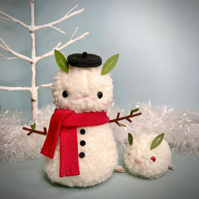 Two fluffy white plush sculptures, one resembles a snowman with a red scarf, black beret hat, twig arms and leaf ears. Next to them is a small fluffy snow bunny with red eyes and leaves for ears.