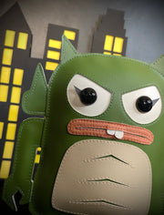 Close up of a vinyl plush of a green Monster of the Black Lagoon-esque character, shaped as a Big Boss Robot figure. Two buck teeth stick out of a closed mouth, with angry eyes. A paper cut out city is in the background.