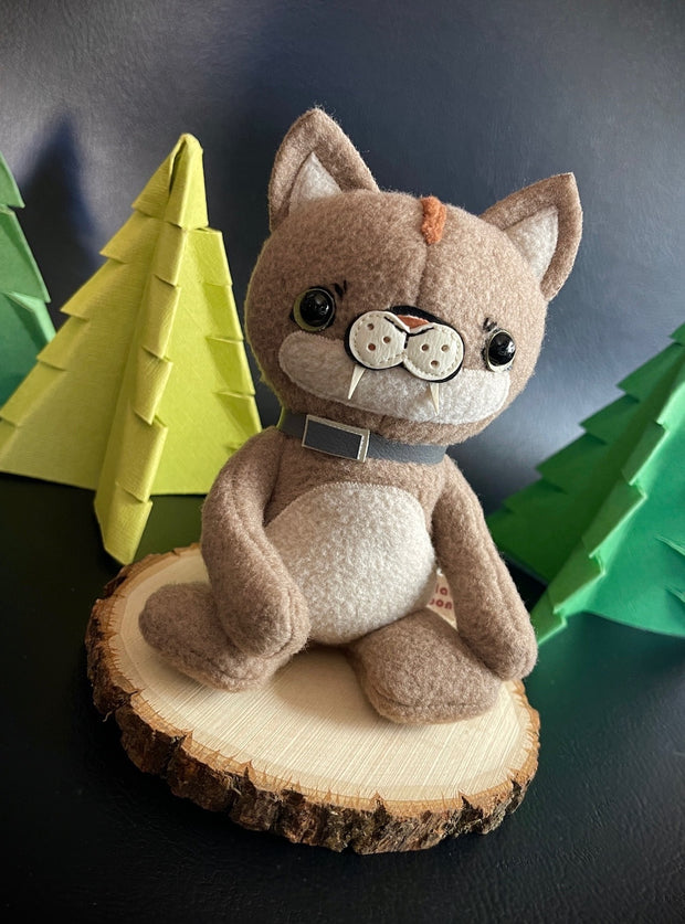 Plush sculpture of a tan puma cub, sitting on a stump with a gray collar around its neck and black bead eyes. In the background are foam pine trees and logs.