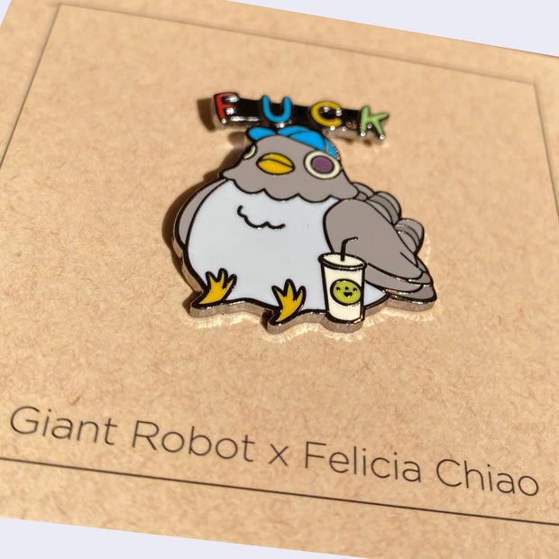 Close up of enamel pin of a chubby, gray pigeon wearing a baseball cap and sitting next to a soda. "Fuck" is written above its head.