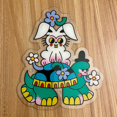 Colorful tattoo style painting on a die cut piece of acrylic on a white bunny with pink color accents and yellow eyes, smiling and riding atop of a serious looking tortoise with flowers on its body, a patterned decorated shell and a black top hat.