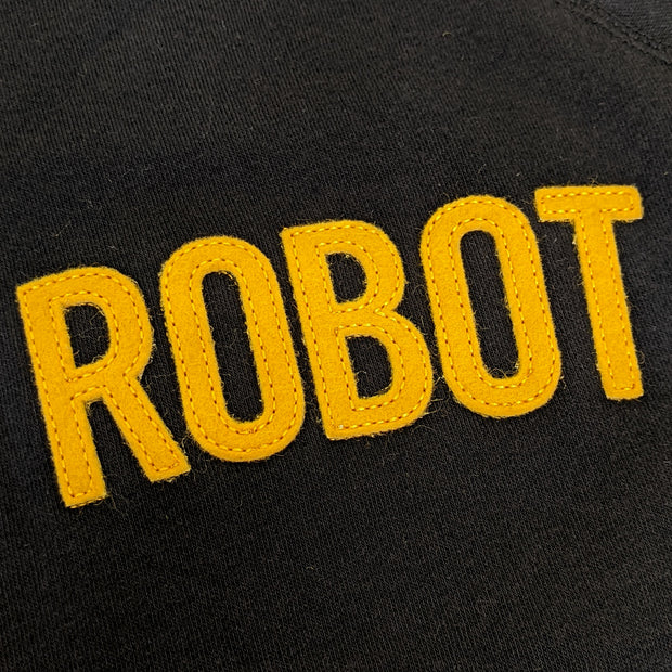 Close up of yellow sewn on felt letters on black sweatshirt, reading "Robot" in all caps.