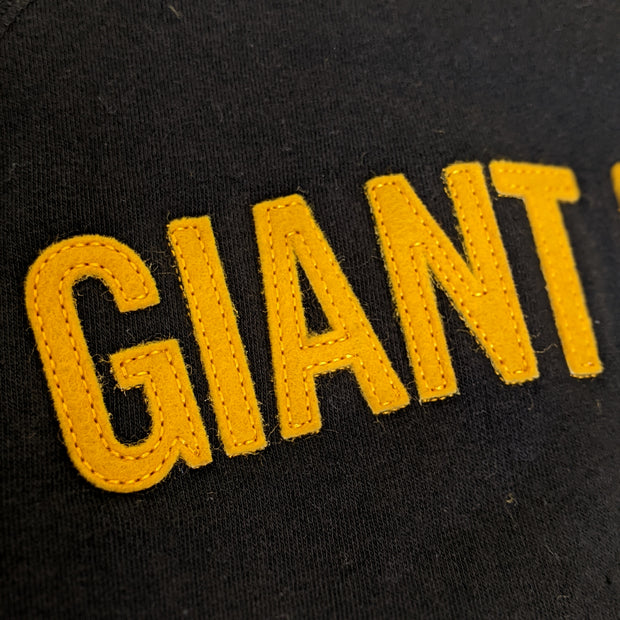Close up of yellow sewn on felt letters on black sweatshirt, reading "Giant" in all caps.