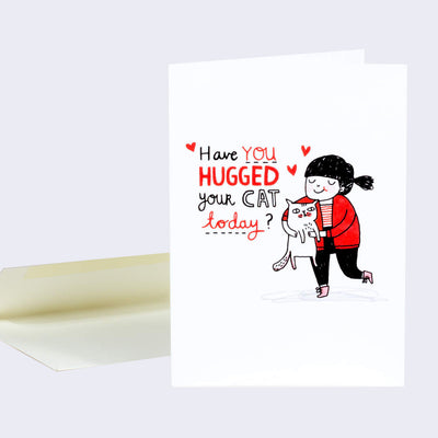 White greeting card with a black and red illustration of a girl with a red jacket and black pants smiling and holding an indifferent looking white cat. "Have you hugged your cat today?" is written in stylized font with red hearts floating nearby.