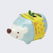 Small ceramic cartoon hedgehog, smiling with a brown nose. Its body is blue and is wrapped in a cardboard packaging sleeve that says "Hedgie Green."