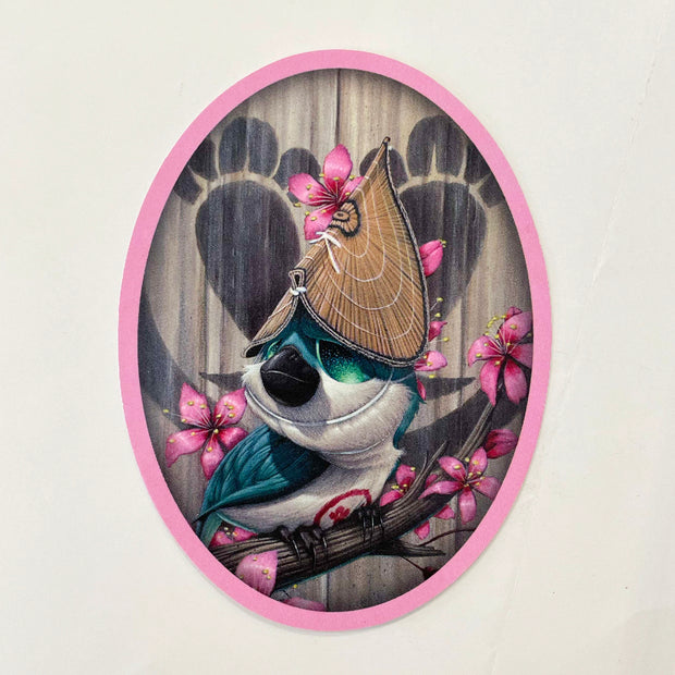 Oval sticker with a pink outline. A small blue illustrated bird with a thick black beak is perched on a cherry blossom tree branch. It has a folded bamboo mat on its hat. 
