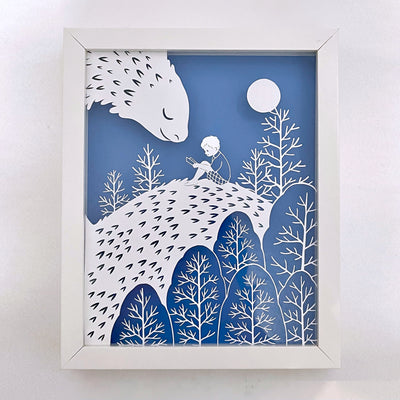 White cut paper illustration on a blue background in a white wooden frame. Cut paper design depicts a young child reading a book, sitting on a kind looking dragon, whose face is turned smiling to the child. A moon and trees are around the two.