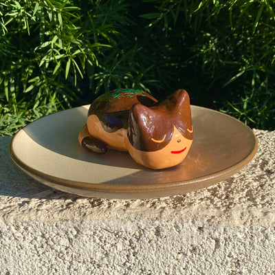 Sculpture of a sleeping tan cat, with simplistic facial features and a red smile. Cat has darker brown accent coloring towards the top of its body, with green flecks on its back to make it resemble a takoyaki.