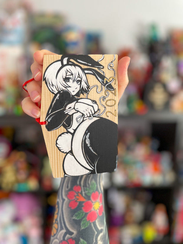 A rectangular wooden token on red string, with illustration of an anime style woman with a curvaceous body and bunny costume lingerie, looking back and exposing her butt that has a cotton bunny tail on it. She wears bunny ears and 2023 is written along the bottom. Painting is black and white.
