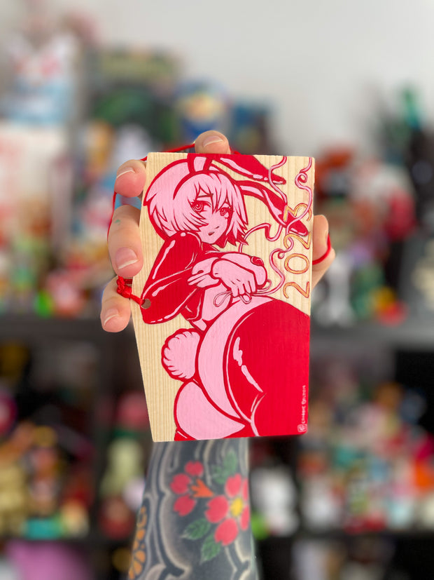 A rectangular wooden token on red string, with illustration of an anime style woman with a curvaceous body and bunny costume lingerie, looking back and exposing her butt that has a cotton bunny tail on it. She wears bunny ears and 2023 is written along the bottom. Painting is pink and red.