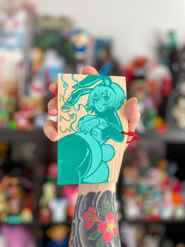 A rectangular wooden token on red string, with illustration of an anime style woman with a curvaceous body and bunny costume lingerie, looking back and exposing her butt that has a cotton bunny tail on it. She wears bunny ears and 2023 is written along the bottom. Painting is light teal and dark teal.