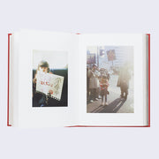 Open two page book spread of photographs with thick white page borders. Left photo is of a little girl holding a large paper with doodles and Japanese writing. Right photo is of a group of people holding up signs, with the girl standing in the middle.