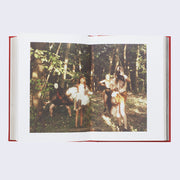 Open two page book spread of a photograph of multiple people standing in the middle of the woods, with sun shining through the leaves onto them.