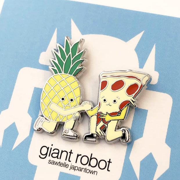 Close up of enamel pin of a pizza and a pineapple, kneeling and clasping hands.