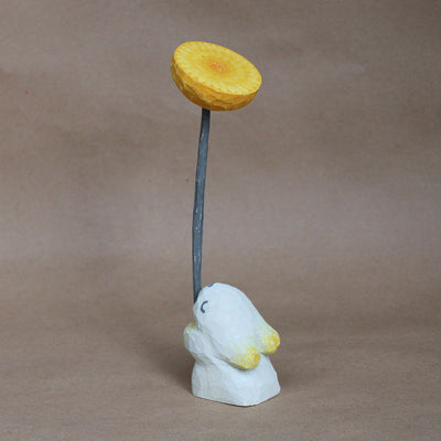 Whittled wooden sculpture of a small white bunny, with eyes closed and very simplistic body, facing away and holding a large whittled wooden yellow dandelion with a long green stem.