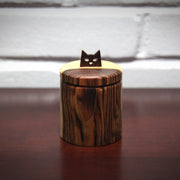 Cylindric wooden box, with a natural wood grain of light wood and darker exterior wood. As a handle for the top is a simplified cat head, with cut out eyes and a small nose.