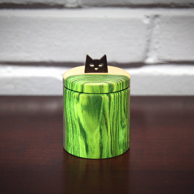 Cylindric wooden box, with a natural wood grain glowing almost neon green from a black light being placed on the bioluminescent wood. As a handle for the top is a simplified cat head, with cut out eyes and a small nose.
