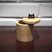 Cylindric wooden box, with a natural wood grain of light wood and darker exterior wood. The lid of the box is pushed slightly off, revealing a container inside. As a handle for the top is a simplified cat head, with cut out eyes and a small nose.