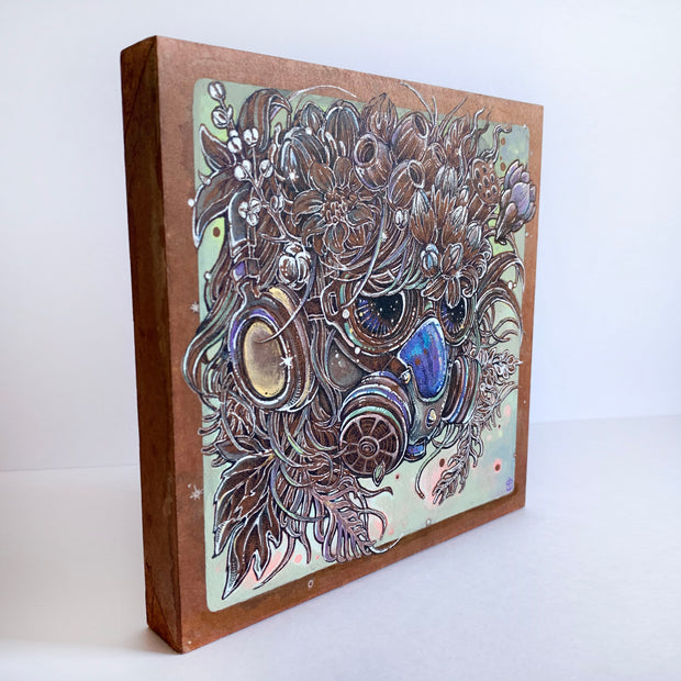 Side angle of  Illustration of a intricate and colorful gas mask, covered all around with wispy hair, flowers and seed pods. Piece is mounted on warm toned wood.