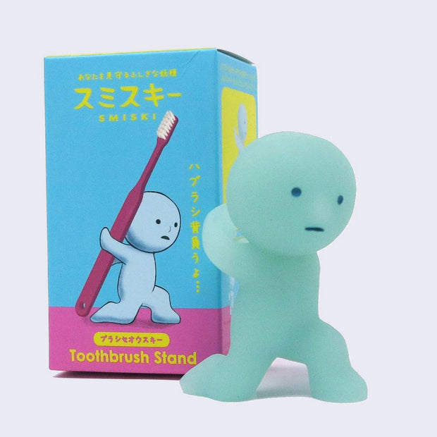 Simplified light teal character kneeling on one knee, with its arms wrapped around an empty place on its back, where one can put a toothbrush. Behind it is its display box.