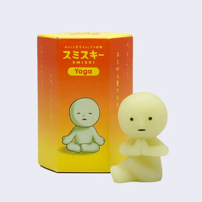 Small simplistic light green/yellow character, with a round head and simple body. It sits in a yoga pose next to its display box.