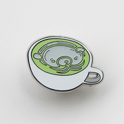 Gif of enamel pin of a matcha latte in a shallow, white mug and a white bear design inside the cup. One image is of the pin in light, the other is of the pin glowing in the dark.