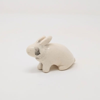 White ceramic rabbit, with simplistic body shapes and no facial features. It is positioned on all 4 paws, slightly sitting back with a gray smudged tear running from where its eye would be. Its ears are slightly back.