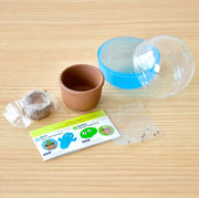 Display of unpacked capsule, showing a packaged disc of peat, a small terra-cotta pot, and empty capsule, seeds and a sheet of instructions.