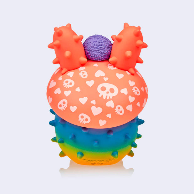 Back of vinyl bunny figure with a rainbow rounded body with spikes like a cacti. Its head is a red mushroom cap with a skull and heart pattern. It has a purple hedgehog on its head.