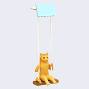 Close up of an orange tabby cat figurine, sitting on a wooden swing attached by cream colored string to a light blue top base where it can rest from a shelf, allowing the cat to freely hang.