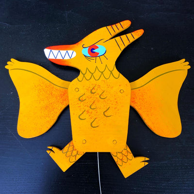 A painted wooden sculpture of Rodan, depicted in a modernish cartoon style. Rodan is a deep yellow with orange splatter accents and a mint blue and red eye, looking to the side with an excited and angry expression, wings and legs spread out