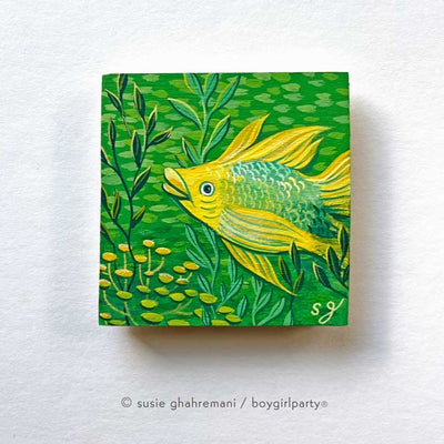 Painting on wooden panel of a yellow fish in a green underwater scene, with lots of simplified fish swimming behind and pieces of kelp nearby.