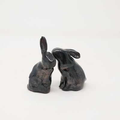 Two black ceramic rabbits with simplistic body shapes and no facial features, one sits on its hind legs with its ears straight up. The other has their ears put back and leans in, appearing to whisper in the other's ear.