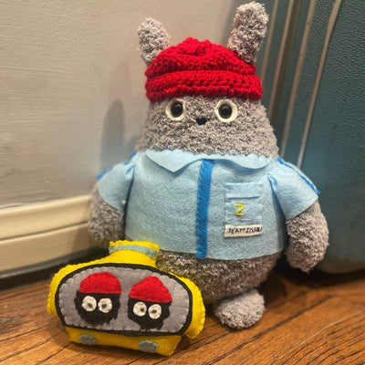 Crocheted cartoon version of Totoro dressed as Bill Murray in The Life Aquatic, with a blue shirt and red beanie. Its accompanied by a yellow submarine type machine with 2 dust sprites in it.