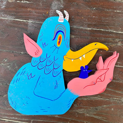 Die cut painted wooden sculpture of a bird monster with white horns, pink ears and pointy teeth with a yellow beak. It holds a small blue mouse in its hand.