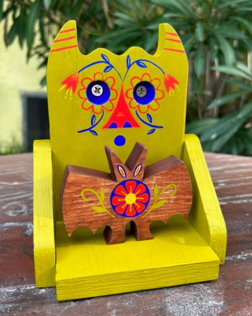 Die cut painted wooden sculpture of an olive green shelf like apparatus that resembles a devil, with flowers painted on its face. It holds a small wooden bat, with a painted flower on its stomach.