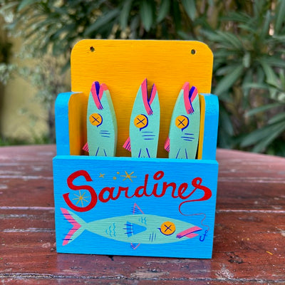 Painted wooden sculpture of a holder that says "sardines" on the front, with an illustration of a mint green sardine. Inside, are three mint green sardines with yellow x'd out eyes.
