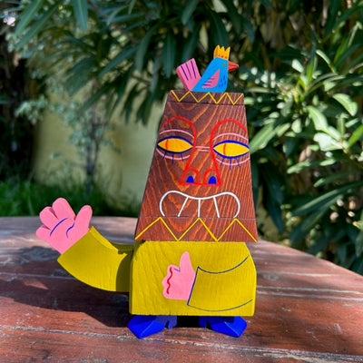 Die cut wooden sculpture of a tiki statue, with a human body. It has one hand extended out and the other as a thumbs up. Atop its head is a blue and pink bird.