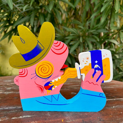 Die cut painted wooden sculpture of a pink man with a green cowboy hat and blue button up shirt. His eyes are yellow with red swirls and he drinks a bottle of yellow liquid with a blue moon label on it.