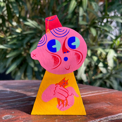 Die cut wooden sculpture of a round headed pink person, with face in whistle pose, wearing a red fez hat. It holds a small orange kitten in its hands.