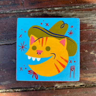 Bright blue painted wooden square, with an illustration of a smiling tabby cat face wearing an olive green cowboy hat.