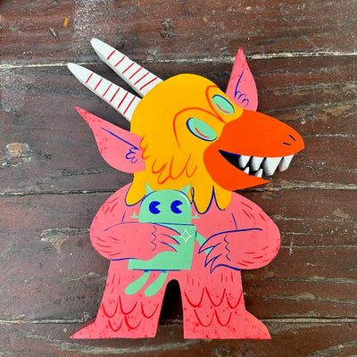 Die cut painted wooden sculpture of a smiling pink monster with a yellow bird face and tall white horns. It holds a small green robot doll between its arms.