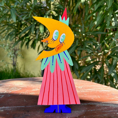 A die cut painted wooden sculpture of a smiling moon, wearing a long pink pin striped cloak with a mint green collar and pointy hat.