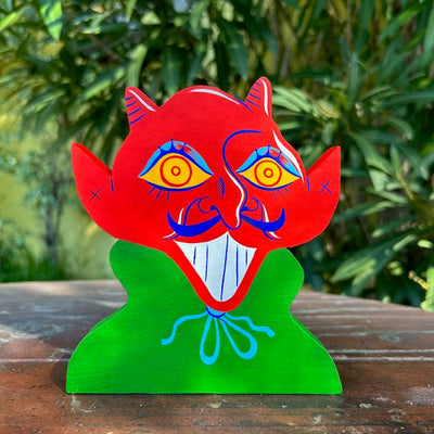 Die cut painted wooden sculpture of a red devil with an upturned mustache, yellow eyes and blue eyeshadow. It wears a green cloak and is only visible from the chest up.