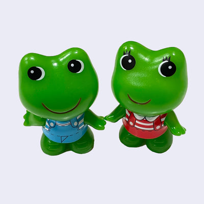 Two small green soft plastic frogs, with large anime style eyes and large simple red smile. They have a large head and small body, with big feet. Koro chan wears a blue set of overalls and Kero Chan has eyelashes and wears a red striped overall set.