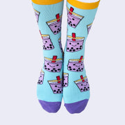 Front view of a model's feet wearing chubby cartoon boba socks. The smiling purple cups are filled with black boba balls. A red straw pokes out.