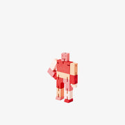 Wooden robot made of square-like shapes, standing with an arm on its hip. It is various shades of pink and red.