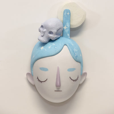 Painted wooden sculpture of a stylized face with closed eyes and blue hair with white sparkles. Atop the head is a purple skull with a full moon.