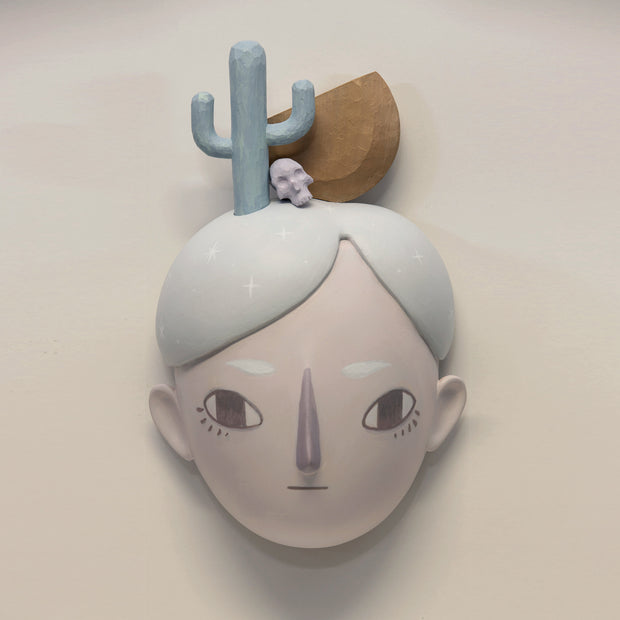 Painted wooden sculpture of a stylized face with light blue hair. Atop their head is a Saguaro cactus, a small skull and a wooden half circle.