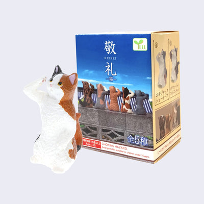 Small plastic cat figure, mainly white with orange and black accent coloring. It stands with its weight on its back paws, looking up and saluting. It stands in front of its product packaging. 